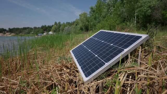 A portable solar panel rests on dry reed grass along a sunny lake shoreline surrounded by green trees. Concept of off-grid power maintenance and renewable energy optimization.