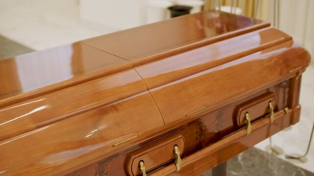 Closed wooden coffin in a bright funeral home or chapel. Solemn farewell ceremony setup with white floral arrangements, empty chairs, and a religious icon.