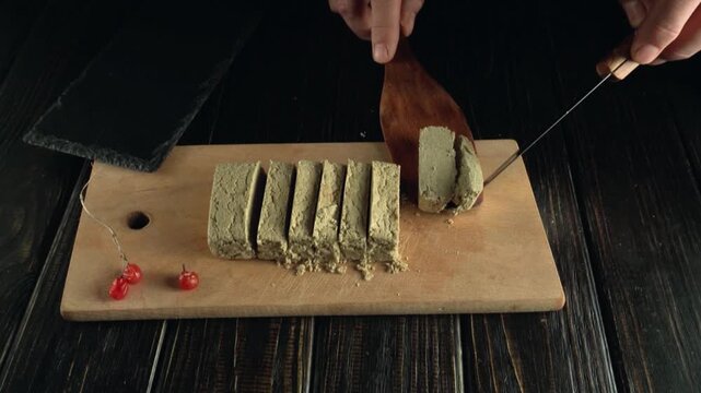 Slicing and serving a block of green herbal paste on a wooden cutting board with a knife and spatula, accompanied by red berries, showcasing the culinary preparation process in a kitchen setting