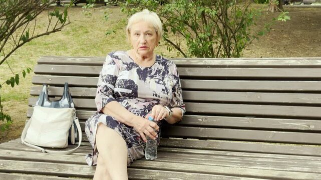An elderly woman talks seriously while holding a water bottle on a park bench. Her concerned expression brings a deeply somber and uneasy feeling.