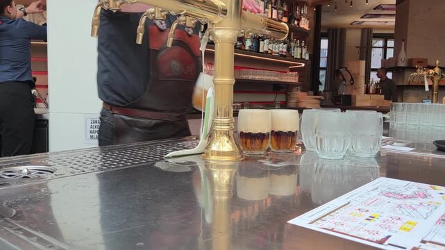 Panning shot of bartender pouring beer from brass tap into glasses on reflective metal bar counter inside pub interior. Beer in Prague