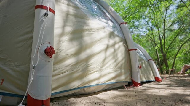 An inflatable camping tent stands fully assembled on sandy ground in a forest campsite. Green trees surround the shelter while sunlight filters through the canopy overhead.