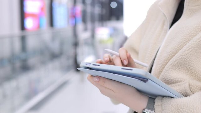 Person holds tablet, uses stylus in public space. Hands visible, wearing beige coat. Background blurred with glass railing. Person focuses on screen, walking forward. Light colors dominate scene