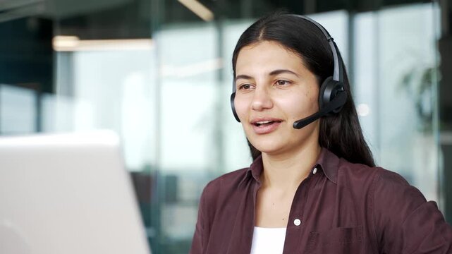 Close up of female customer service operator in headset working on laptop and talking to client in modern office. Woman support agent provides assistance, answers questions and helps solve issues.