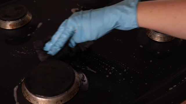 Scrubbing dirty gas stove with soapy foam closeup, hand in glove wiping black metal burner area, kitchen chore routine