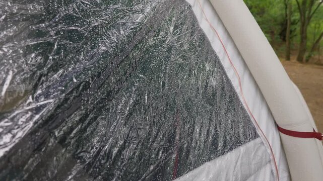 Raindrops cover the transparent plastic window of a camping tent pitched in a wet forest. Concept of weather protection, waterproof shelter, and outdoor survival during heavy rain.