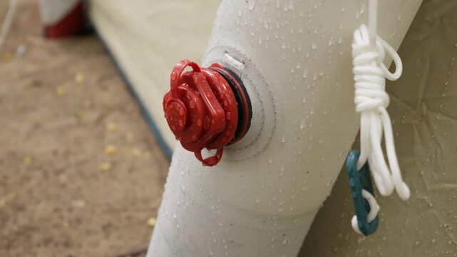 A detailed close-up shows the red inflation air valve of an inflatable camping tent covered with rain water droplets. A white guyline rope is neatly tied adjacent to the beam outdoors.