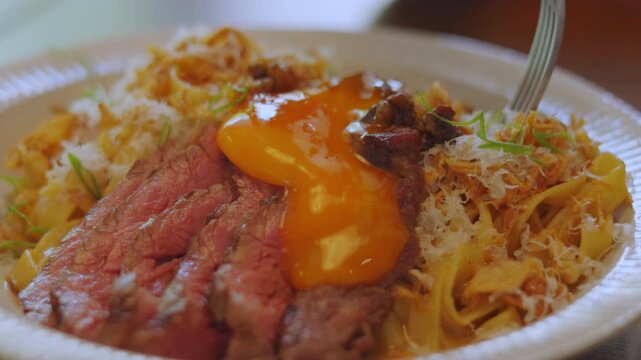 Close up of using a fork to twirl delicious pasta coated in oozing egg yolk and served with sliced medium-rare beef steak. Appetizing gourmet dining and mouth-watering culinary food concept.