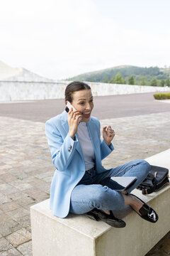 Businesswoman celebrating success by phone call, sitting outdoors with tablet and bag, copy space