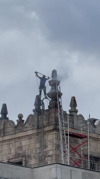 Worker cleaning decorative architectural elements on the rooftop of a historic building in Warsaw, Poland. Maintenance of historic architecture and urban building exterior against a modern city skylin