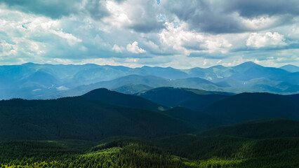 Fototapeta premium Hazy mountain tiers stretch to the horizon with alternating bands of light and shadow. Blue tones soften the scene while nearby forests form a textured foreground of spruce.