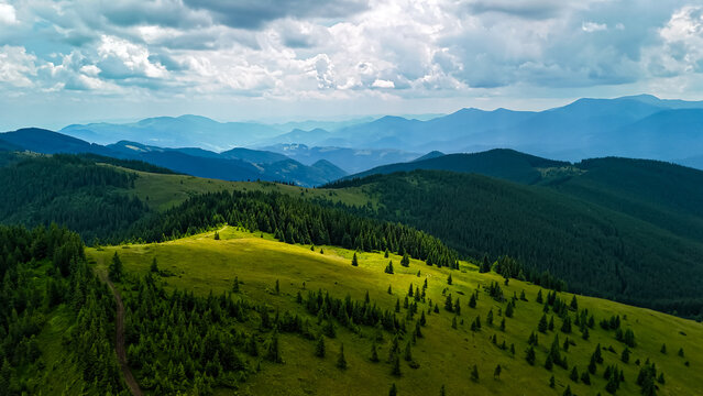 Curving ridge path leads through sunny grass toward a realm of folded green mountains. Puffy clouds spread across the vault of sky and light dances gently on the landscape.