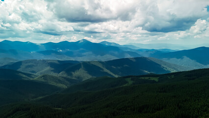 Fototapeta premium Vast mountain range with rolling forested hills under dramatic clouds and soft sunlight. Blue haze layers fade to a distant horizon. Peaceful wilderness vista evokes adventure and calm.