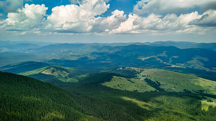 Fototapeta premium Rolling ranges drift beneath a bright sky as forests and meadows share the slopes. The aerial scene captures distance depth and light that shifts across a timeless mountain realm.