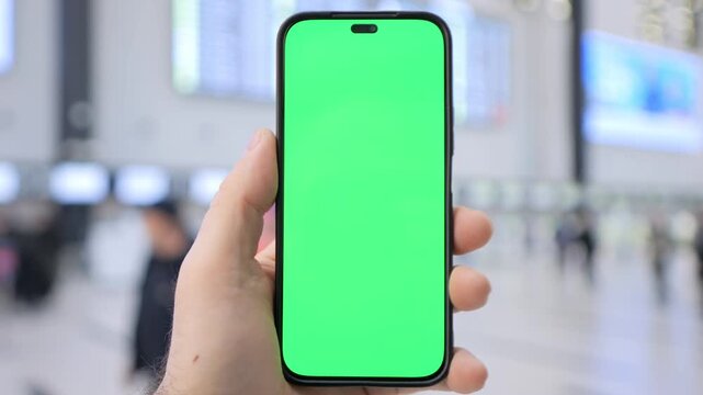 Hand holds phone with green screen in busy station. Blurred crowd moves in background. Useful for app demos or travel-related content. Ideal for digital marketing or UI mockups