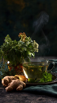 A steaming cup of herbal tea, garnished with fresh ginger and herbs, on a dark, textured base