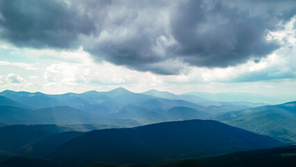 Fototapeta premium Dark cloud bases hover above a sea of blue mountains where narrow valleys gleam with light. The fore ridge lies in shadow giving depth and mood to this airy and remote terrain