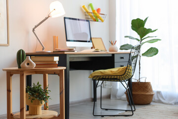 Fototapeta premium Wooden side table, desk with computer, lamp and chair near white wall in home office, selective focus. Interior design