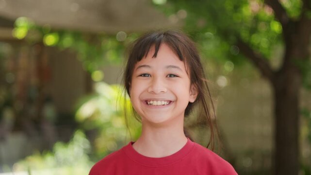 Portrait of adorable Asian kid looking directly at the camera with a gentle smiling face in a red shirt standing outdoors with beautiful green bokeh background. Childhood happiness lifestyle concept.