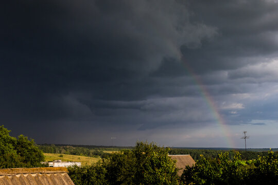 Brooding storm clouds sweep over open countryside as a delicate rainbow appears in the brightening gap above sunlit trees and roofs the dramatic sky captures the moment between fury and peace