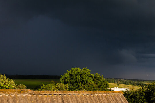 A towering storm front looms over bright green fields and trees as sunlight grazes the landscape while the sky turns nearly black creating a powerful contrast that signals heavy rain ahead