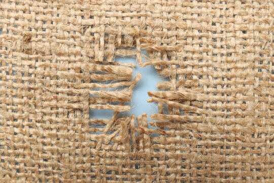 Burlap fabric with torn hole on light blue background, top view