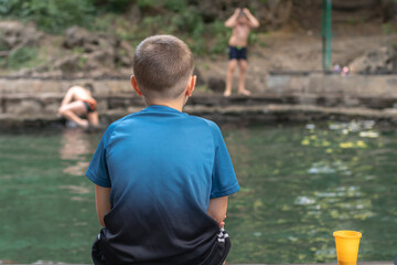 Fototapeta premium A boy sitting on the edge of a pond and watching other children swimming.