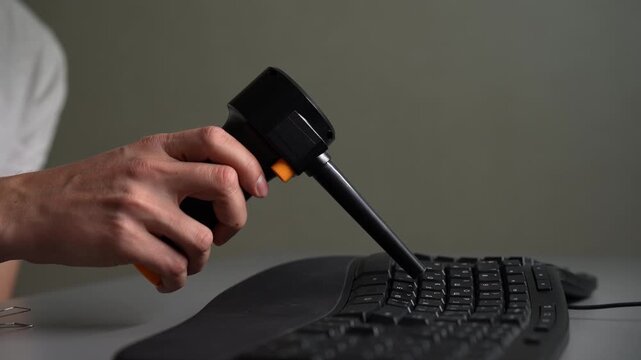 Professional technician using cordless electric air duster to blow dust and debris from between keys of black computer keyboard, ensuring proper electronic maintenance and office hygiene, slow motion.