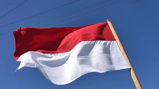 Indonesian flag (Sang Merah Putih) waving on a bamboo pole against a clear blue sky background.