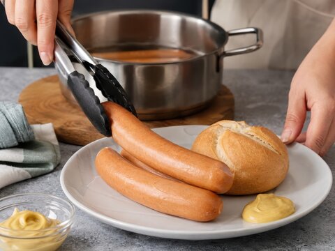 Bockwurst anrichten mit H&auml;nden &ndash; Frisch gegarte Wurst wird mit Zange auf einem Teller neben Br&ouml;tchen und Senf serviert. Authentische Zubereitung eines beliebten deutschen Imbissklassikers.