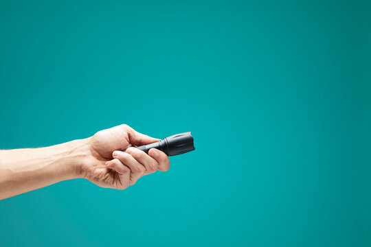 Hand holding black flashlight with bright led light on a teal green background, illuminating and showing the way
