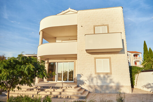 Modern three-story villa with natural stone facade, curved balconies, and sliding glass doors under clear sky