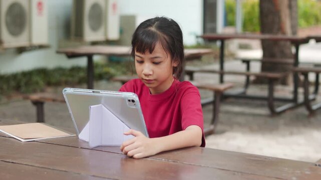 Asian kid using stylus pen on a digital tablet for e-learning at outdoor school table. Selective focus shows the young student engaging with technology for modern education and digital study concepts.