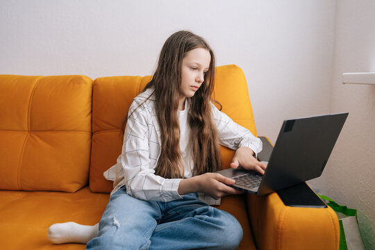 Young girl focused on studies and leisure, sitting on yellow sofa at home typing on laptop, enjoying online learning, browsing and digital connectivity in casual, comfortable setting.