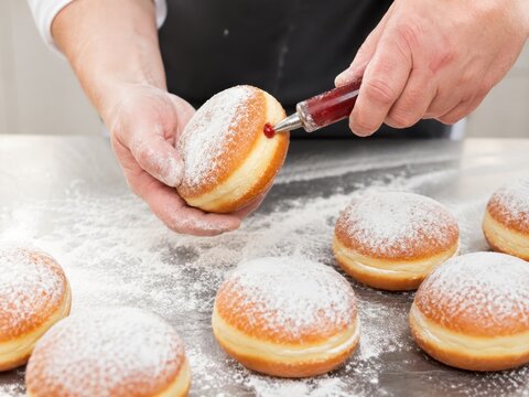 Berliner mit H&auml;nden f&uuml;llen &ndash; Marmelade wird in frisch gebackene, mit Puderzucker best&auml;ubte Berliner gespritzt. Authentische Nahaufnahme klassischer deutscher Backkunst in der K&uuml;che.