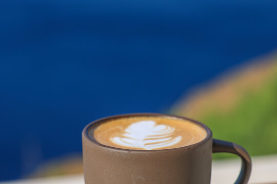 A cup of latte with leaf‑pattern foam rests on a terrace overlooking the Aegean Sea in Greece, blending morning calm with Mediterranean simplicity.
