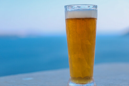 A tall glass of golden beer stands against the blue Aegean Sea in Greece, glowing in sunlight and evoking Mediterranean leisure and calm.
