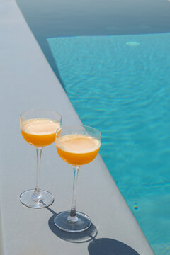 Two orange cocktails rest beside a turquoise pool under the Greek sun, evoking Mediterranean leisure and bright summer refreshment.