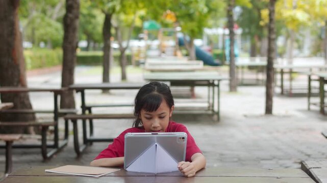 Asian kid doing homework on digital tablet at outdoor campus table. The selective focus shows the student enjoying active e-learning, highlighting modern education technology and tech-savvy childhood.