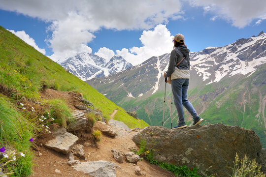 Female hiker with trekking poles looking at high rocky mountains with snowy peaks under blue sky.