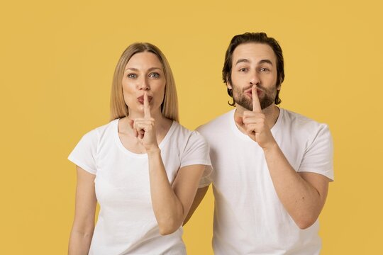 Focused Caucasian Male And Female Making Shushing Gesture With Finger On Lips, In Playful Manner, Against Bright Yellow Background. Ideal For Communication, Silence Or Secrecy Concept.