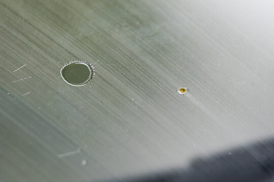Macro of water on a brushed metallic surface with a circular droplet ring and tiny pit. Linear grain and soft gradients create a technical abstract with subtle depth.
