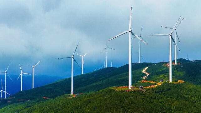 Wind turbines standing on green mountain ridge generating renewable clean energy under cloudy sky in Guilin, China.