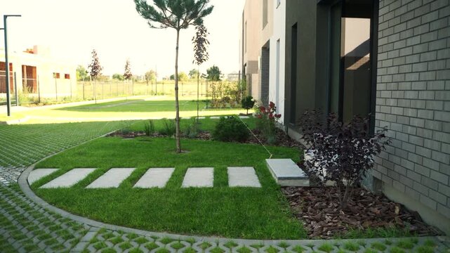Modern house backyard with a lush green lawn, concrete slab stepping stone path, young pine tree, and turf block paving on a sunny day revealing the building's exterior