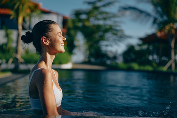 Fototapeta premium Smiling woman in a white swimsuit enjoys a sunny poolside moment, hair in a neat bun. Bright outdoor light highlights a relaxed lifestyle, wellness, and self care beside clear water on a tropical