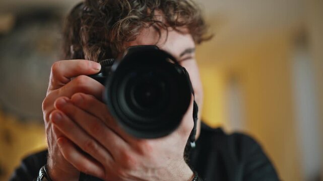 Young man aiming DSLR camera at viewer close up. Photographer shooting indoors.