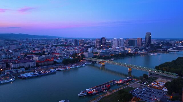 Broad aerial perspective of Bratislava and Old Bridge. Urban lights glow across the riverfront promenade and modern business district at dusk.