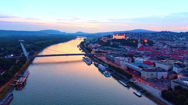 Aerial view of Bratislava Castle and Most SNP at sunset. Drone shot over Danube river showing illuminated city buildings, castle hill, suspension bridge, evening sky.