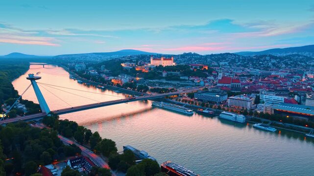 Aerial vista of Most SNP bridge and historic Bratislava. A pink twilight sky reflects on the river surface flowing through the city center.