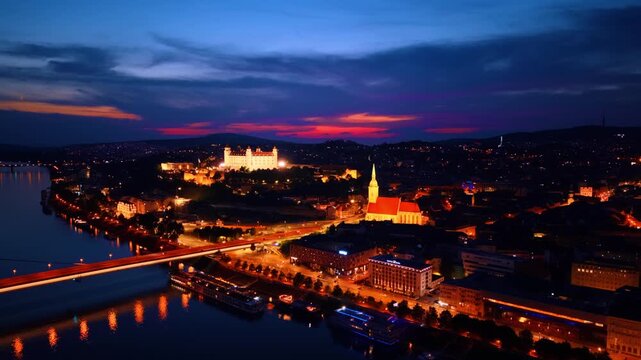 Night aerial view of illuminated Bratislava Castle and old town. High angle shot of lit castle on hill, cathedral, riverbank road, dark sky with sunset horizon.
